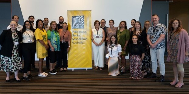 A large group of conference delegates pose around a pull-up banner displaying the Indigenous Dental Association Australia logo